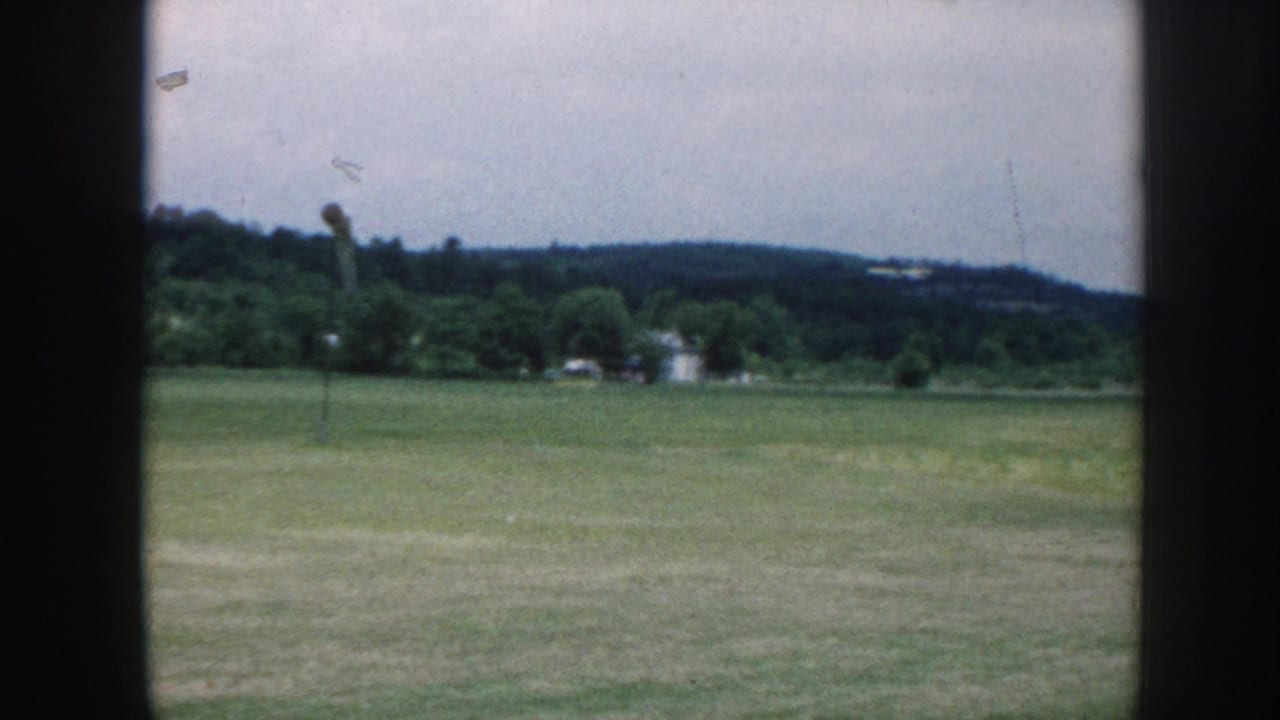 Rural grass airfield viewed through a window, c. 1958 Nebraska — flat green turf, wooded ridgeline, overcast sky, wind poles at field edge.