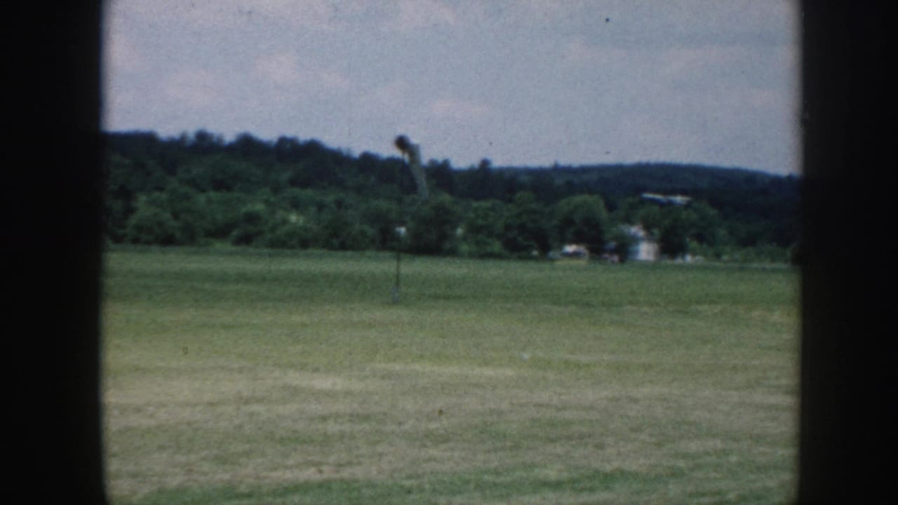 Rural grass airfield framed by window mullions, c. 1958 Nebraska — wide flat turf, wooded ridge, overcast sky, wind pole at right.