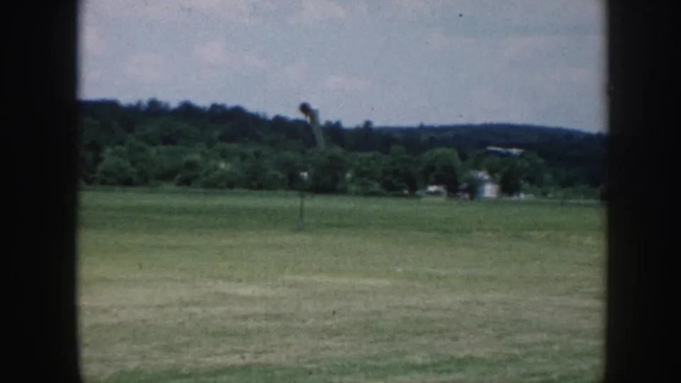 Rural grass airfield framed by window mullions, c. 1958 Nebraska — wide flat turf, wooded ridge, overcast sky, wind pole at right.