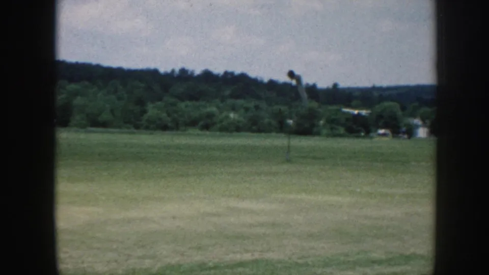 Small single-engine aircraft with white fuselage and blue tail landing on a grass strip, c. 1958 Nebraska — window-framed observer perspective.