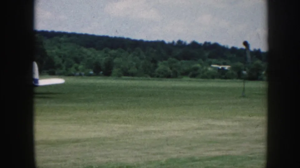 Small single-engine aircraft with blue tail rolling on grass strip after landing, c. 1958 Nebraska — framed by window dividers, wooded ridge beyond.