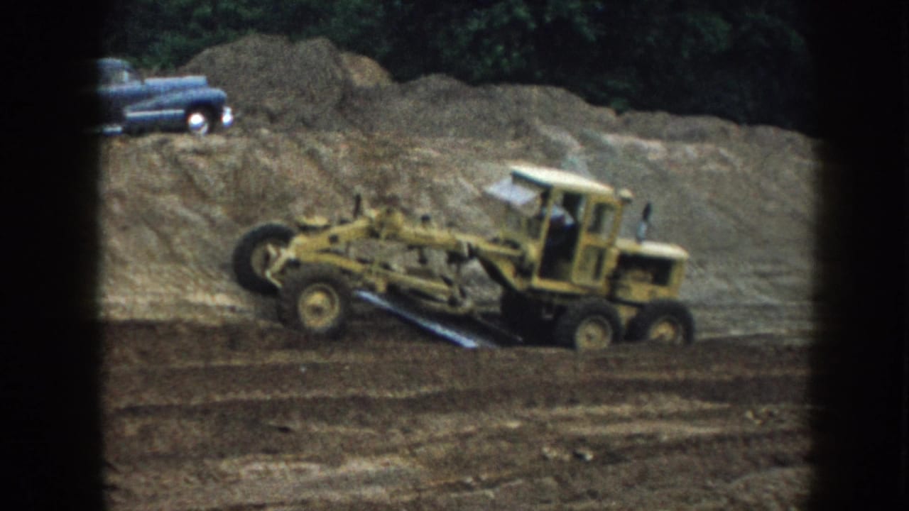 Yellow motor grader at work on a dirt construction site, Nebraska 1958; blue American sedan parked on earthen berm behind, 8mm frame vignetting at edges.