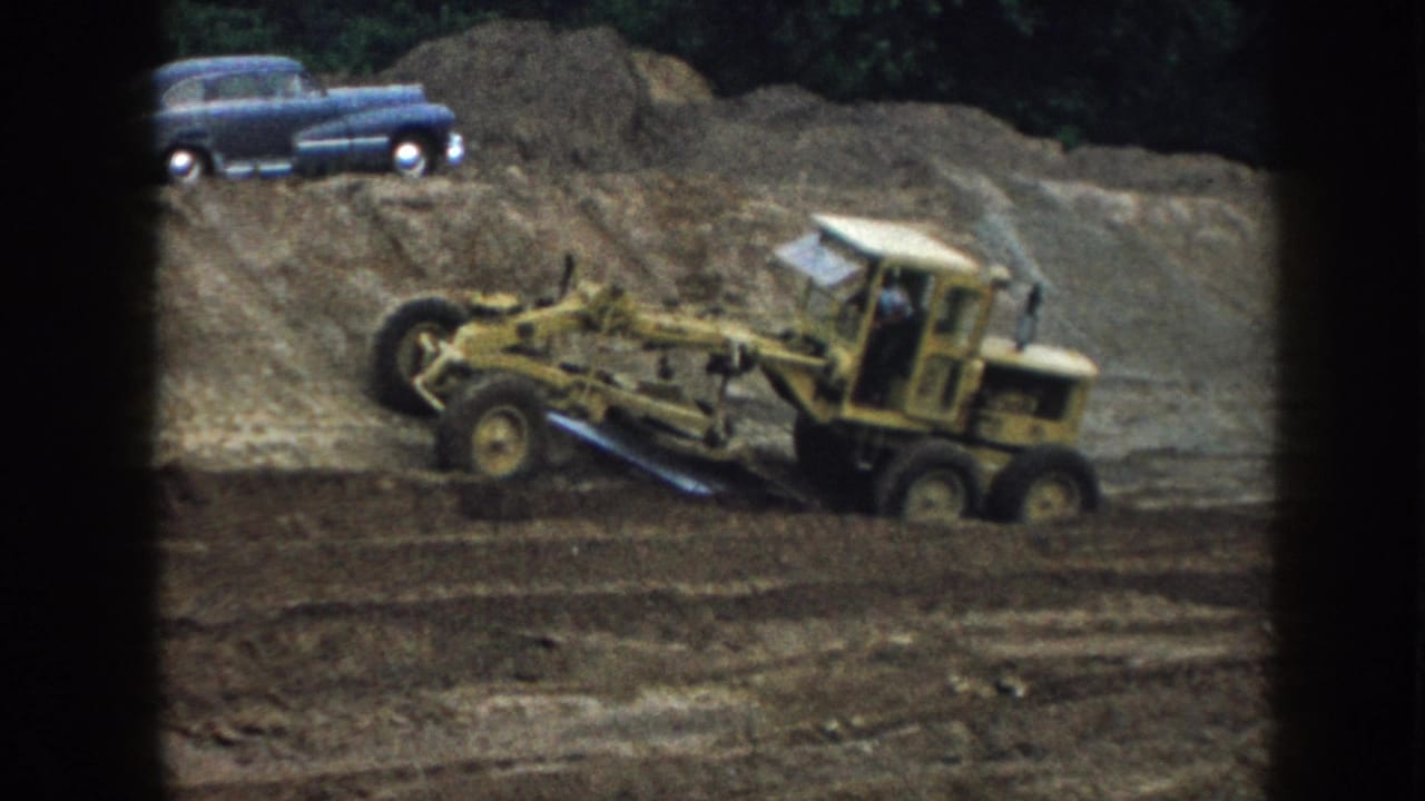 Yellow motor grader moving across graded earth, blue classic sedan parked in upper-right background among trees.