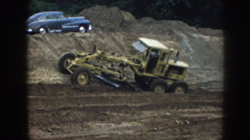 Low-angle 8mm view of yellow motor grader blade assembly working graded earth, Nebraska 1958; blue classic sedan parked on earthen berm at upper left.