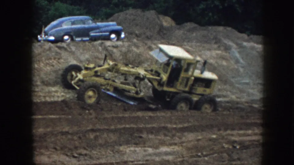 Rear-quarter view of yellow motor grader on graded dirt, Nebraska 1958; blue classic American sedan visible on earthen berm, warm 8mm color saturation.