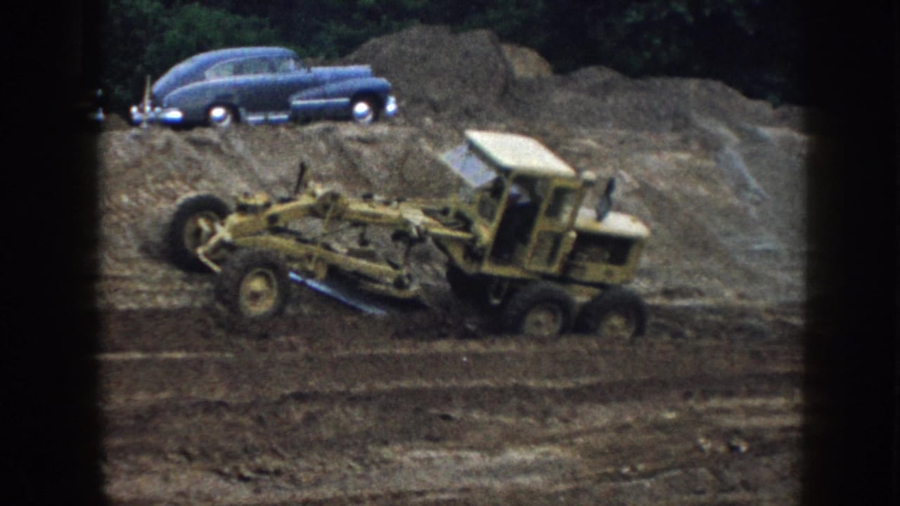 Yellow road grader working across a graded dirt surface, dark blue 1950s sedan parked on an earth mound in the background.