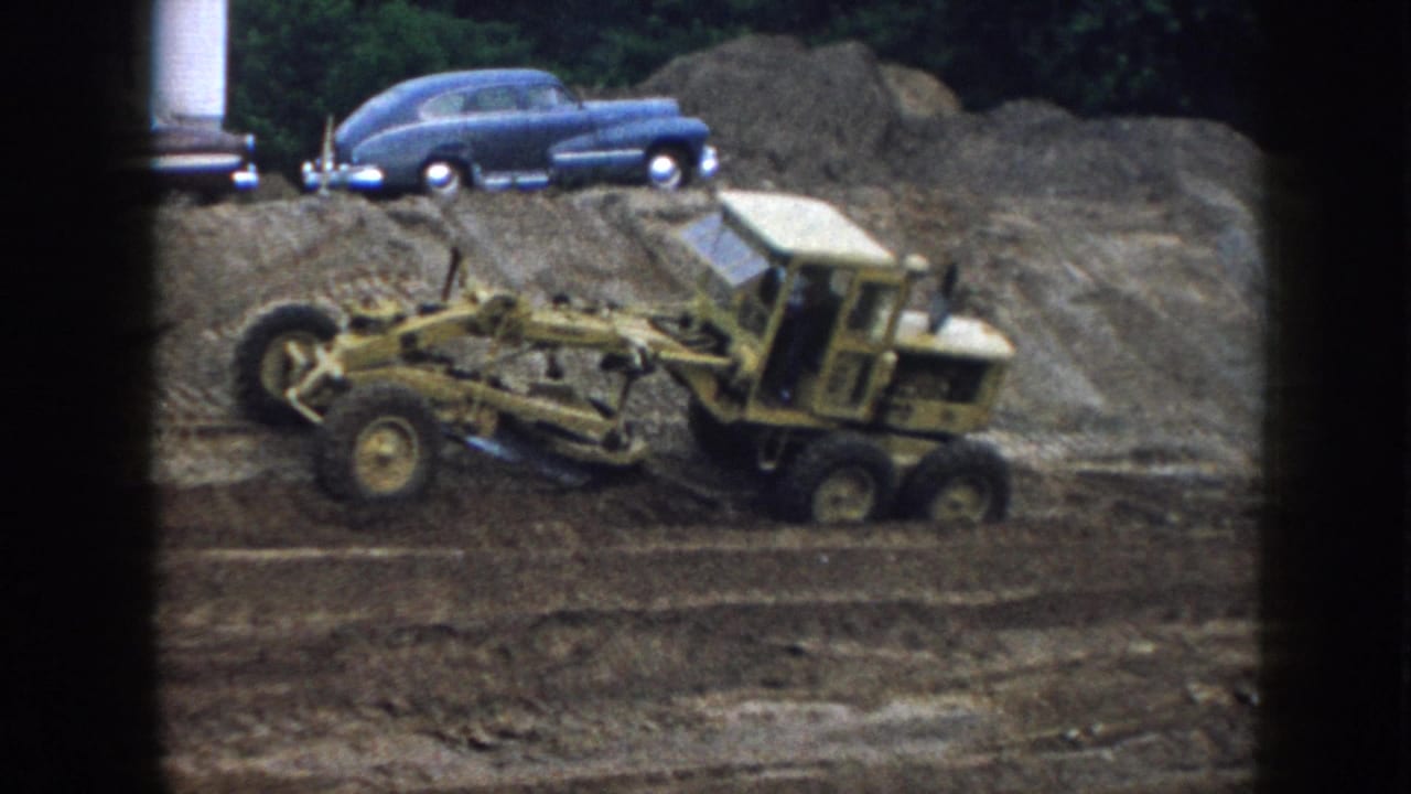 Yellow road grader in motion across dirt, dark blue vintage sedan visible on an elevated earth mound in the upper right.