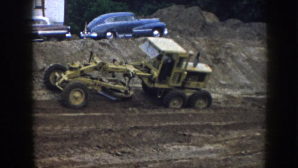 Close-angle view of a yellow motor grader at a Nebraska earthwork site, circa 1958; a dark blue sedan and possibly a second vehicle rest on a raised earth mound.