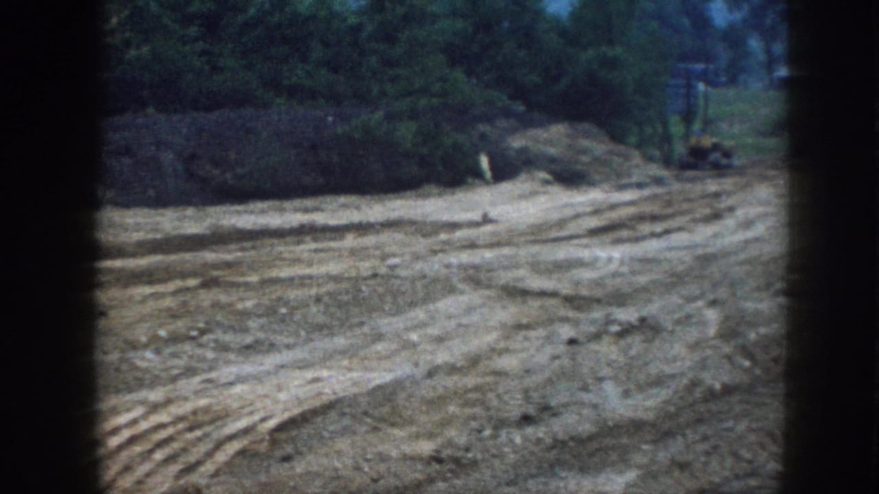 1958 Nebraska rural construction site on 8mm: freshly graded bare earth with machinery tracks, red farm tractor at distance, tree line under overcast sky.