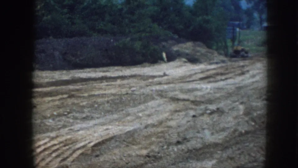 1958 Nebraska rural construction site on 8mm: freshly graded bare earth with machinery tracks, red farm tractor at distance, tree line under overcast sky.