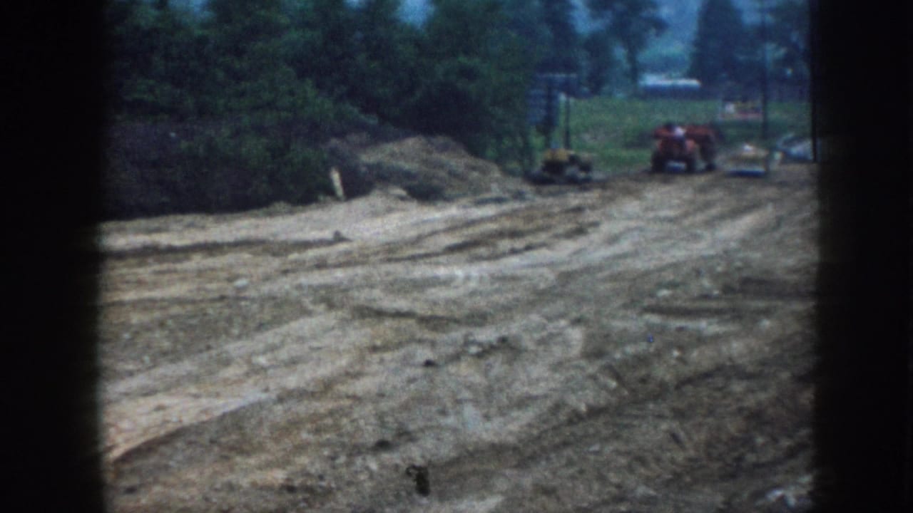 8mm Nebraska construction footage c.1958: wide bare earth terrain with equipment tracks, red tractor at distance, overcast tree line and film-gate border visible.