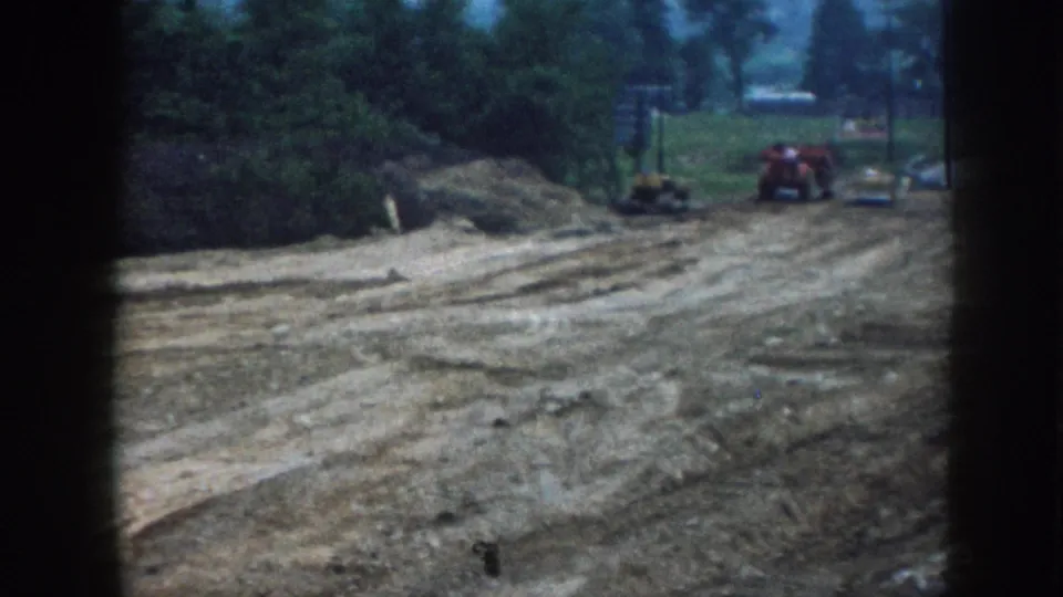 8mm Nebraska construction footage c.1958: wide bare earth terrain with equipment tracks, red tractor at distance, overcast tree line and film-gate border visible.