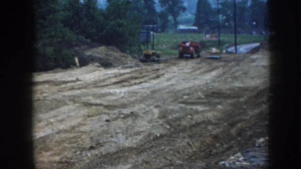 1958 Nebraska grading site on 8mm: red tractor and small yellow bulldozer at mid-ground, utility poles and green tree line visible, bare earth in foreground.