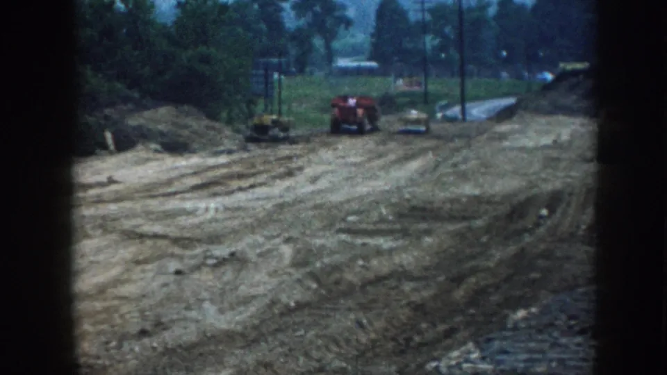 8mm archival footage, Nebraska 1958: red tractor and yellow bulldozer on graded site, utility poles and tree line under diffuse overcast light, film-gate border visible.