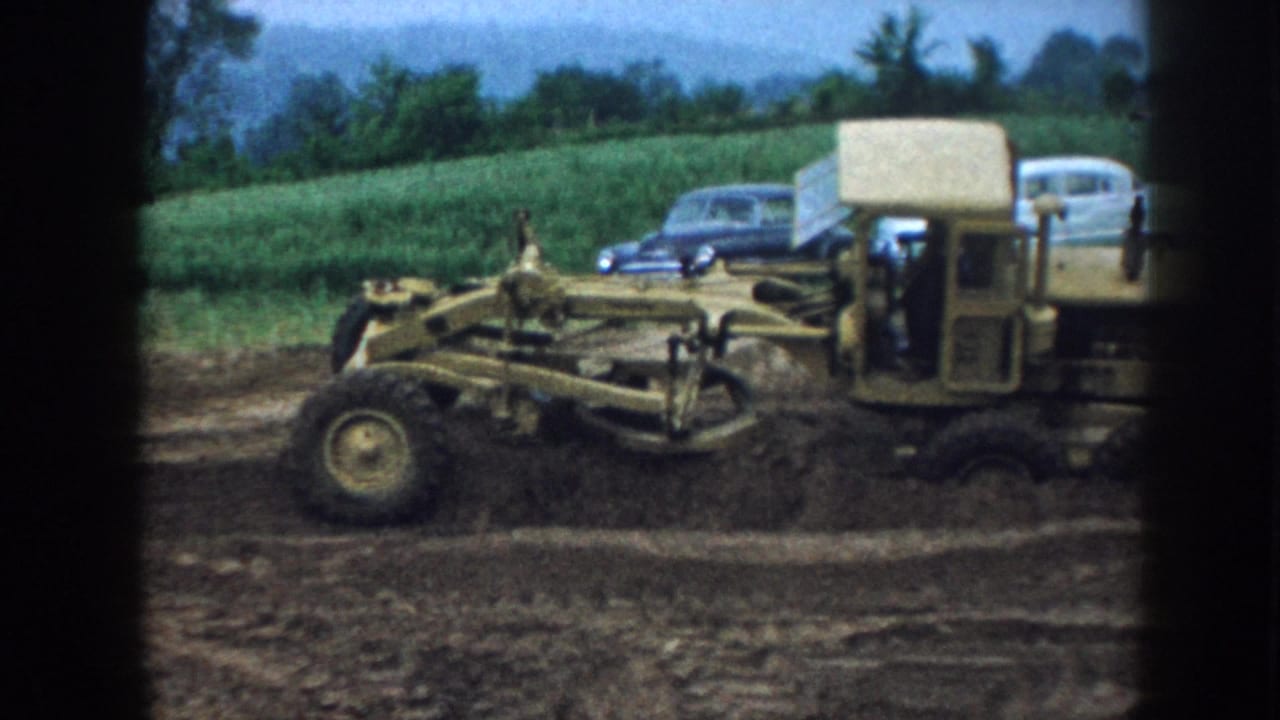 Yellow motor grader grading a rural ditch in Nebraska, 1958; mid-century American sedans parked behind green crop rows.