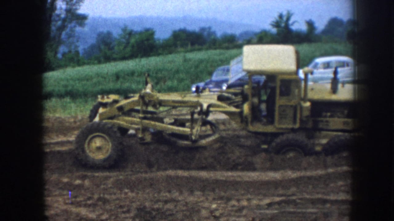 Motor grader passes along a rural ditch in 1958 Nebraska; period automobiles and green crop fields frame the background.