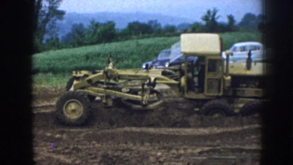 Motor grader passes along a rural ditch in 1958 Nebraska; period automobiles and green crop fields frame the background.