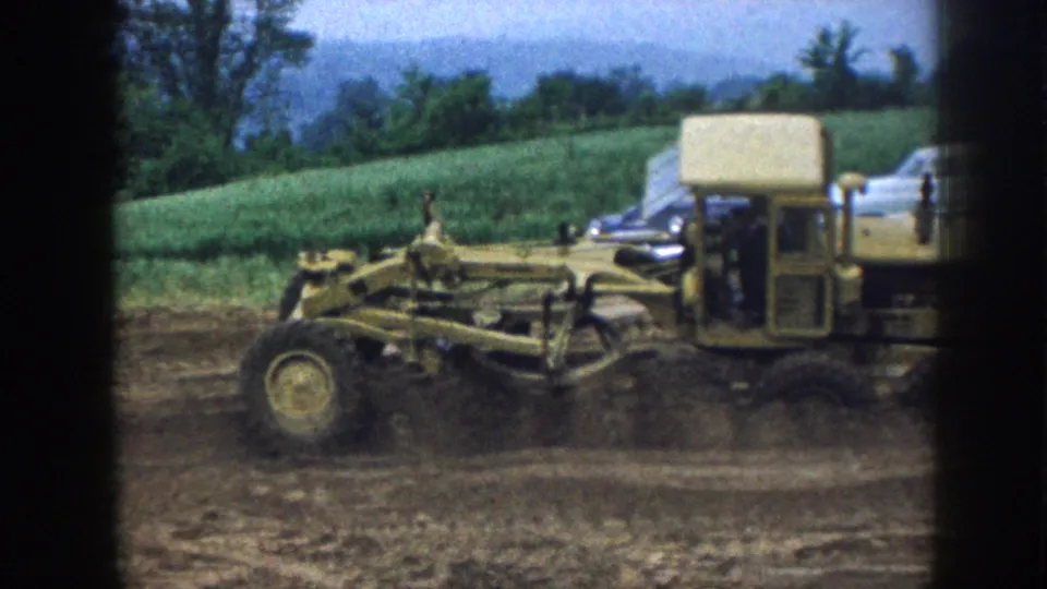 Mid-century yellow road grader grading a rural ditch in Nebraska, 1958; overcast sky and cultivated hillside visible behind.