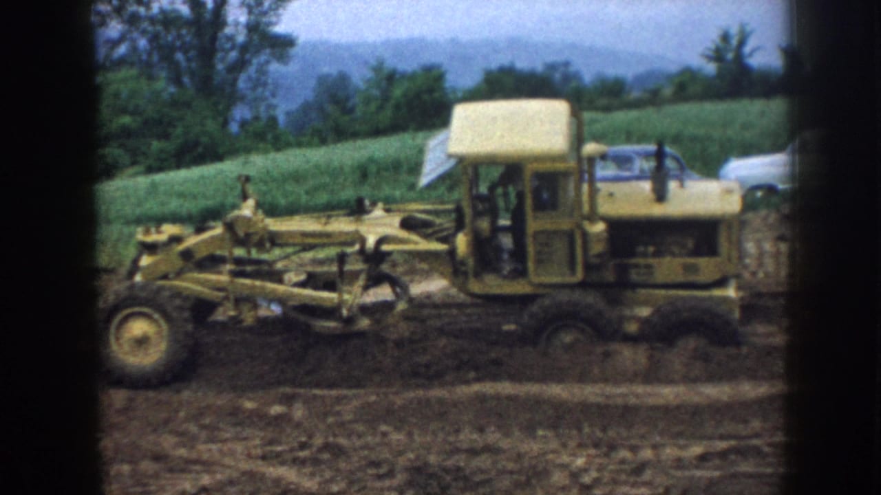 Yellow motor grader working on a dirt construction site with green crop fields and trees behind