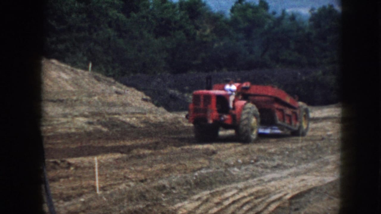 Red tractor-scraper on a graded dirt slope with an evergreen tree line behind