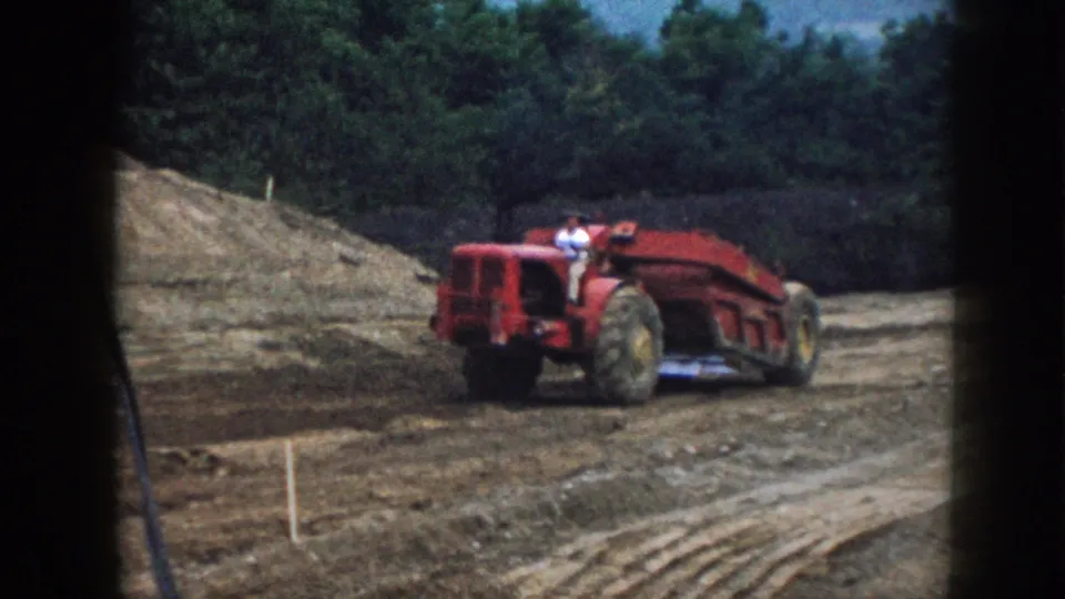 Red tractor-scraper with a visible operator in light clothing working graded terrain at a rural Nebraska construction site, circa 1958; survey stakes and dark film borders visible