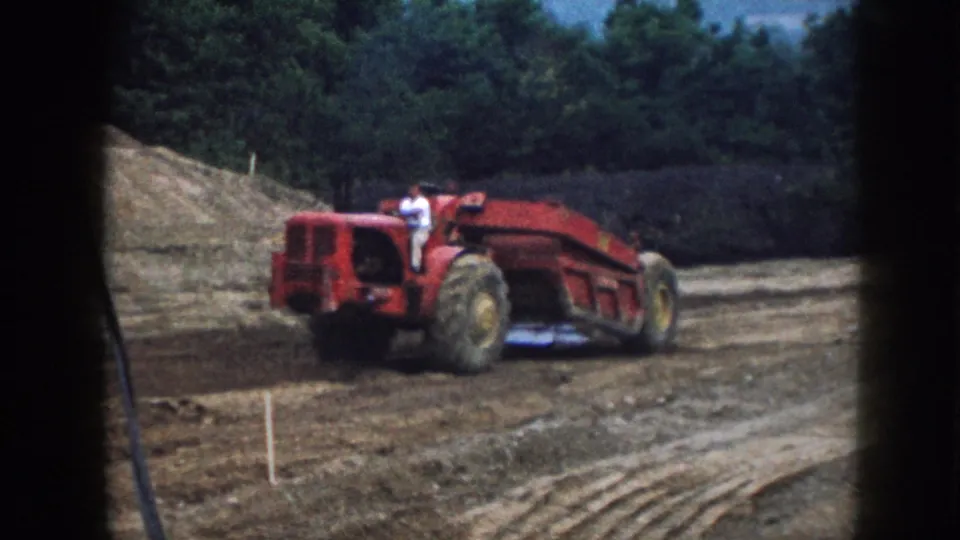 Red tractor-scraper with operator in light clothing traversing graded earth at a rural Nebraska construction site, circa 1958; survey stakes visible in the foreground