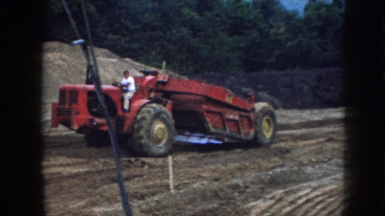 Rear view of red earthmoving scraper on a dirt grading site; operator in white shirt at controls; earthen mound and conifers behind.