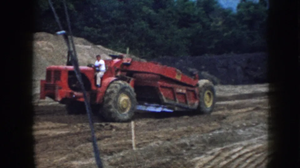 Rear view of red earthmoving scraper operating on a graded site, Nebraska, circa 1958; bowl attachment raised; earthen mound and conifer treeline in background.