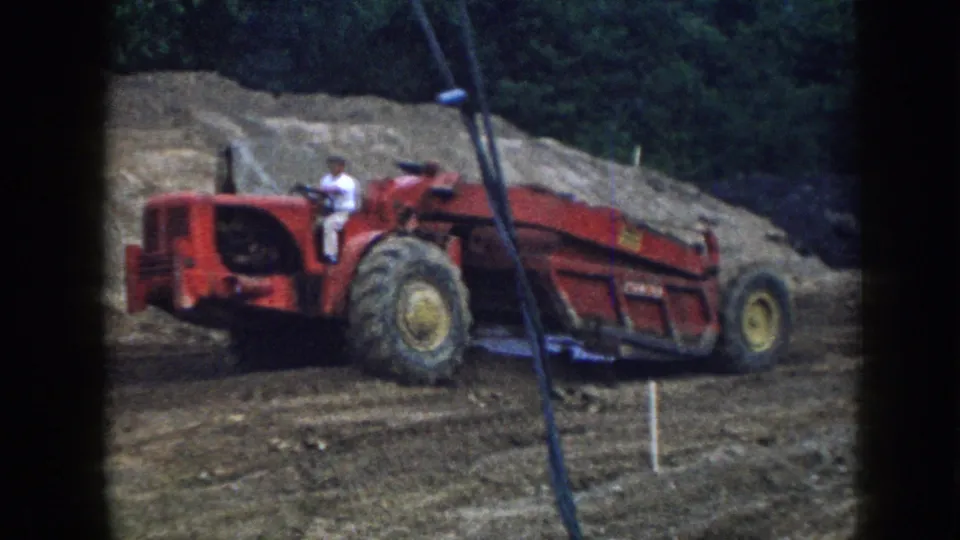Red earthmoving scraper repositioning on a graded construction site, Nebraska, circa 1958; operator in white shirt; survey stakes and earthen mound visible.