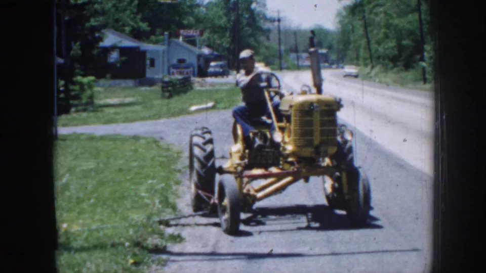 Yellow vintage farm tractor driven on a rural road, Nebraska, circa 1958; male operator in cap; small rural buildings and roadside trees visible behind.