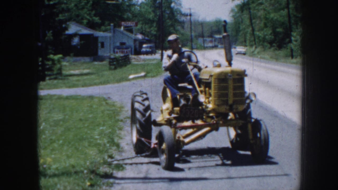 Adult male in work clothes and cap pilots a yellow vintage tractor along a rural Nebraska road, circa 1958, with buildings and green trees behind.