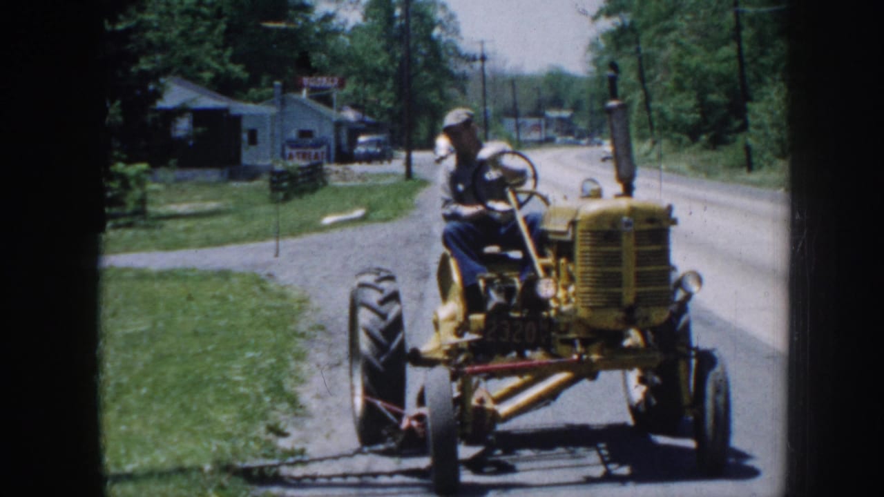 Man on yellow tractor approaching camera on rural road, utility pole and commercial buildings with signage visible behind.