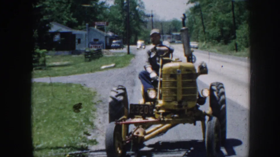 Yellow vintage tractor with a front number plate or model badge approaching camera head-on, rural Nebraska circa 1958, operator in cap visible at the wheel.