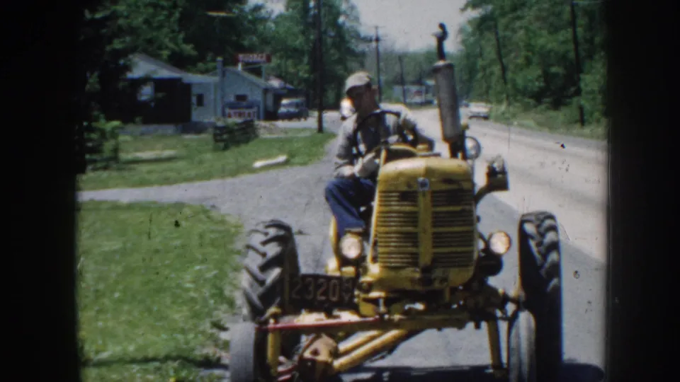 Yellow vintage tractor at close range with operator in cap; front plate or model number visible; small commercial buildings and parked cars in rural Nebraska background, circa 1958.