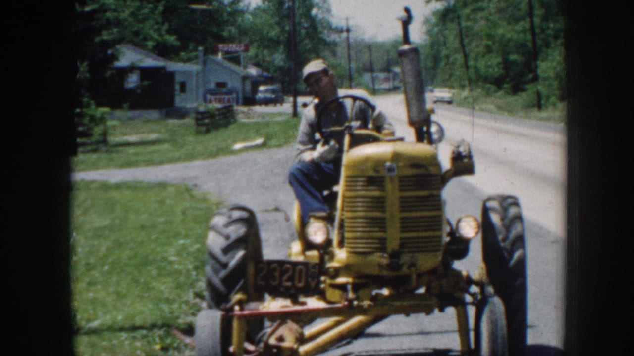 Older man in work cap driving yellow tractor down a rural road, small commercial buildings and trees visible behind.