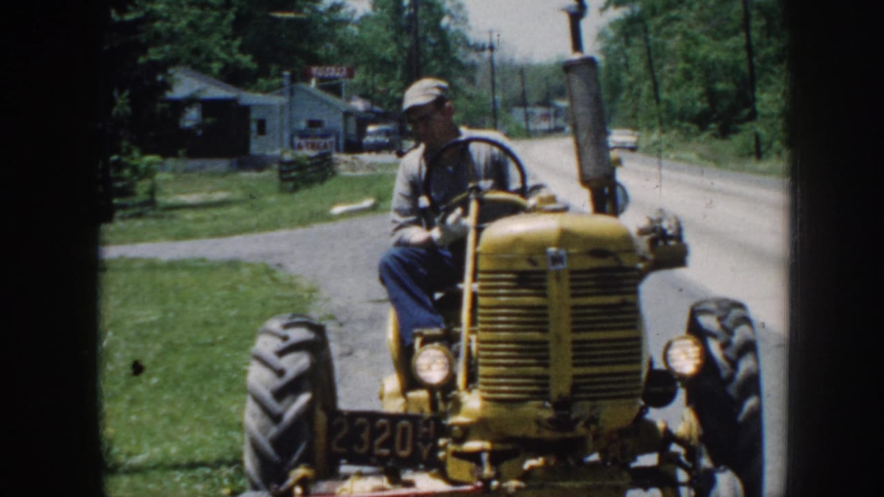 Man in cap and glasses on yellow tractor viewed from roadside; rural buildings and trees in background.