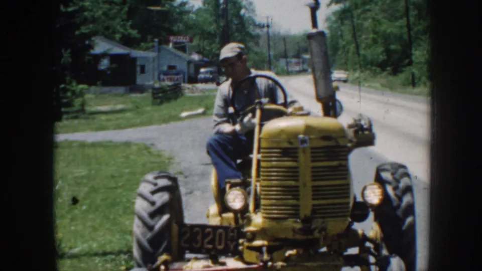 Roadside view of older man steering yellow tractor; period commercial buildings visible behind, consistent with late-1950s rural Nebraska.