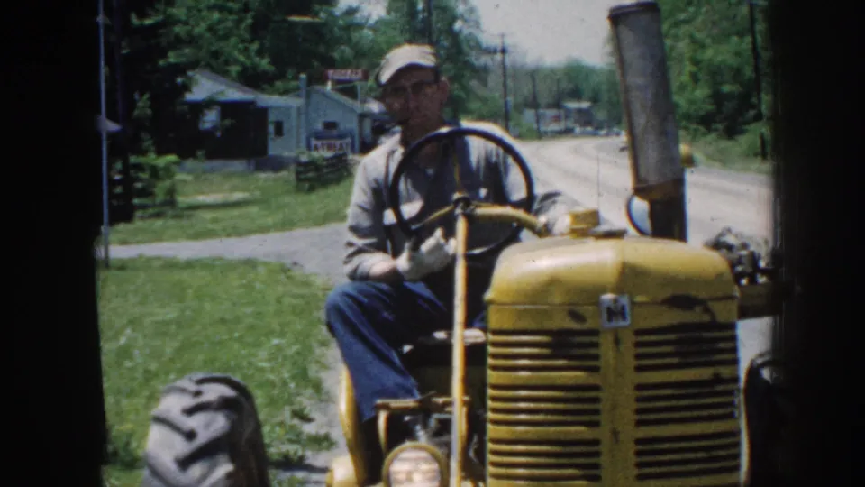 Close-up of yellow tractor front showing International Harvester badging; driver visible above; rural summer foliage soft in background.