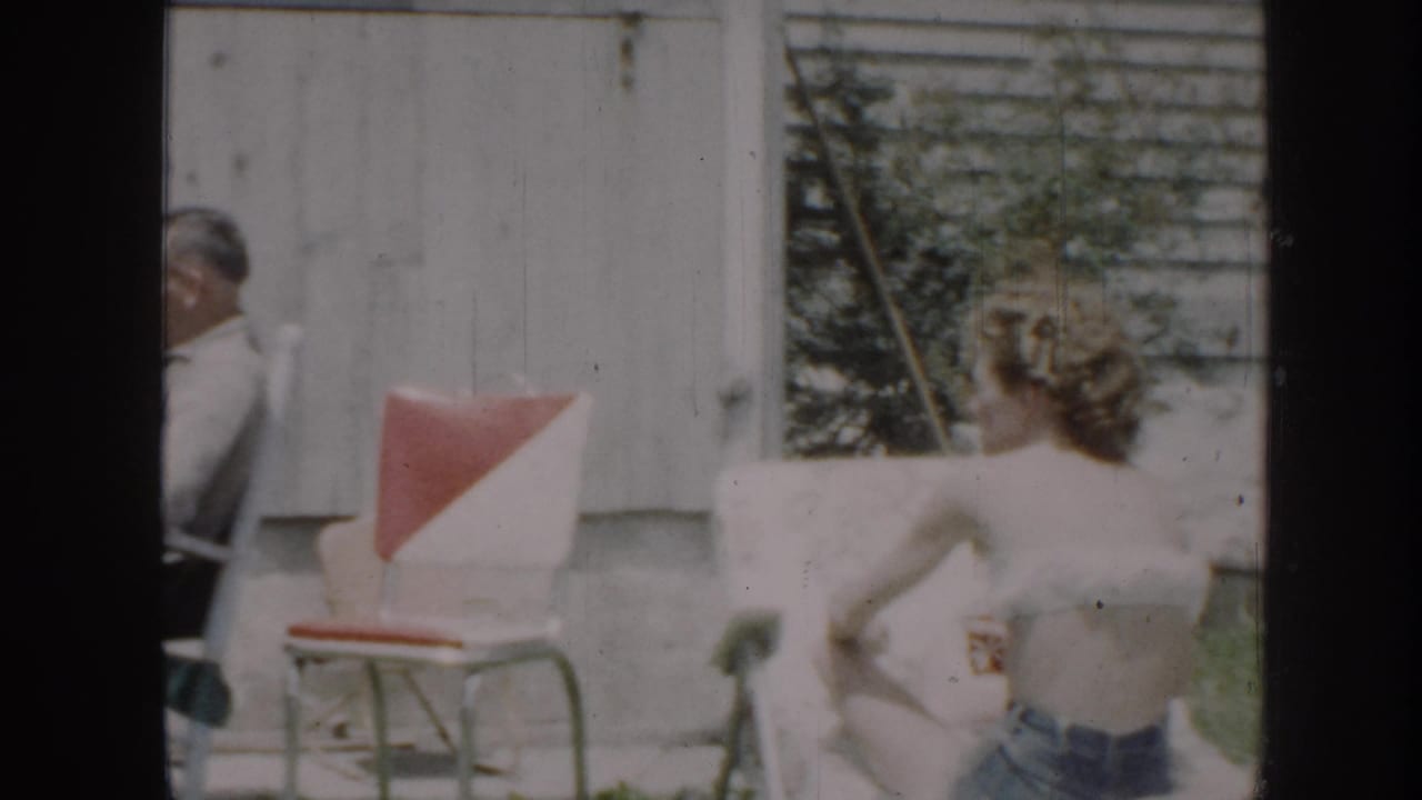 1950s home movie frame: woman in denim shorts and white top, back to camera, in a sunlit backyard with red-cushioned metal lawn chair and outbuilding visible.