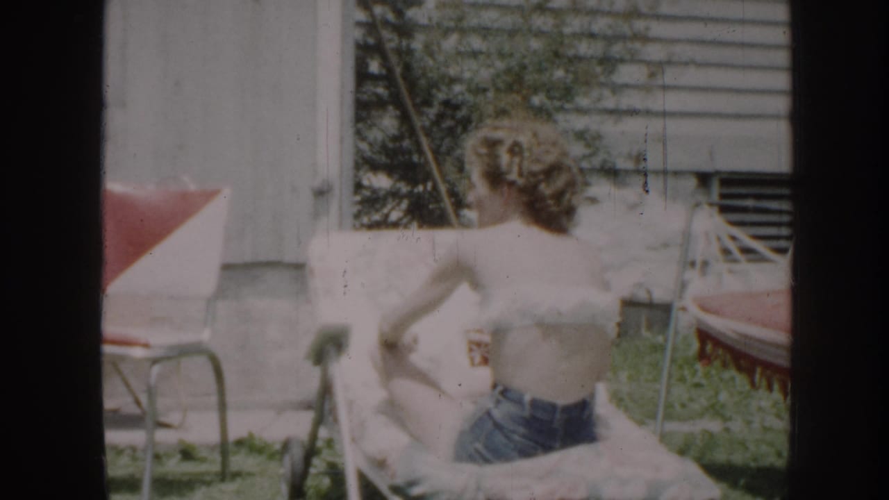 Woman from behind in white top and denim shorts near a red-cushioned lawn chair, outbuilding and trees in the background.