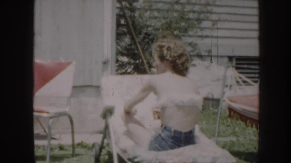 Late 1950s 8mm home movie, Nebraska: woman in denim shorts viewed from behind beside a red-cushioned metal lawn chair in a bright sunlit yard.