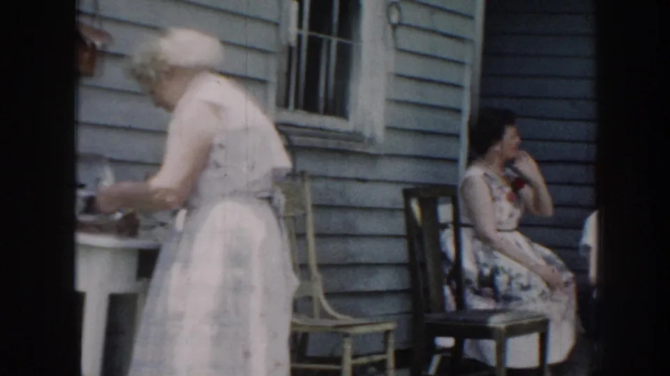 1950s porch gathering, Nebraska: older woman handles dishes at a small table while a younger woman sits in a wooden chair; light blue clapboard siding and window behind them.