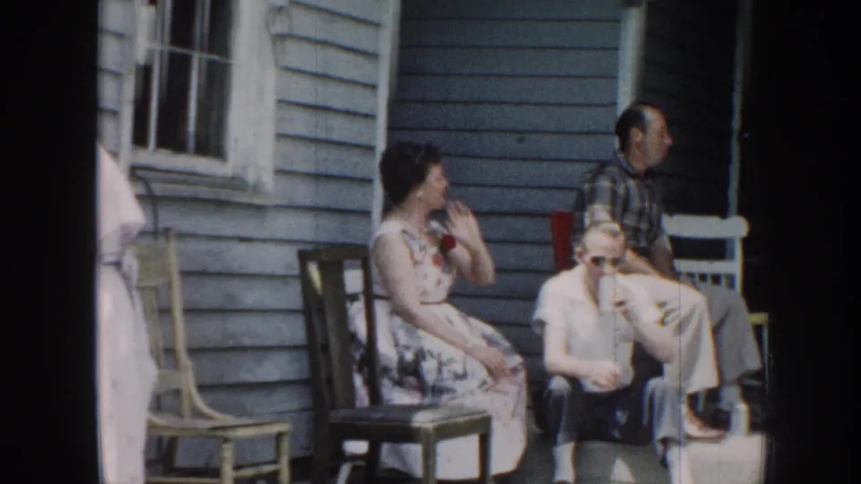 Late 1950s Nebraska porch scene: woman in floral dress seated in wooden chair, man in plaid shirt at her side, additional guests visible against light blue clapboard siding.