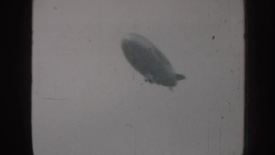 Blimp with gondola silhouetted against washed-out 8mm sky, appearing larger and more defined than the previous frame; consistent with a 1950s American airship passing low overhead.