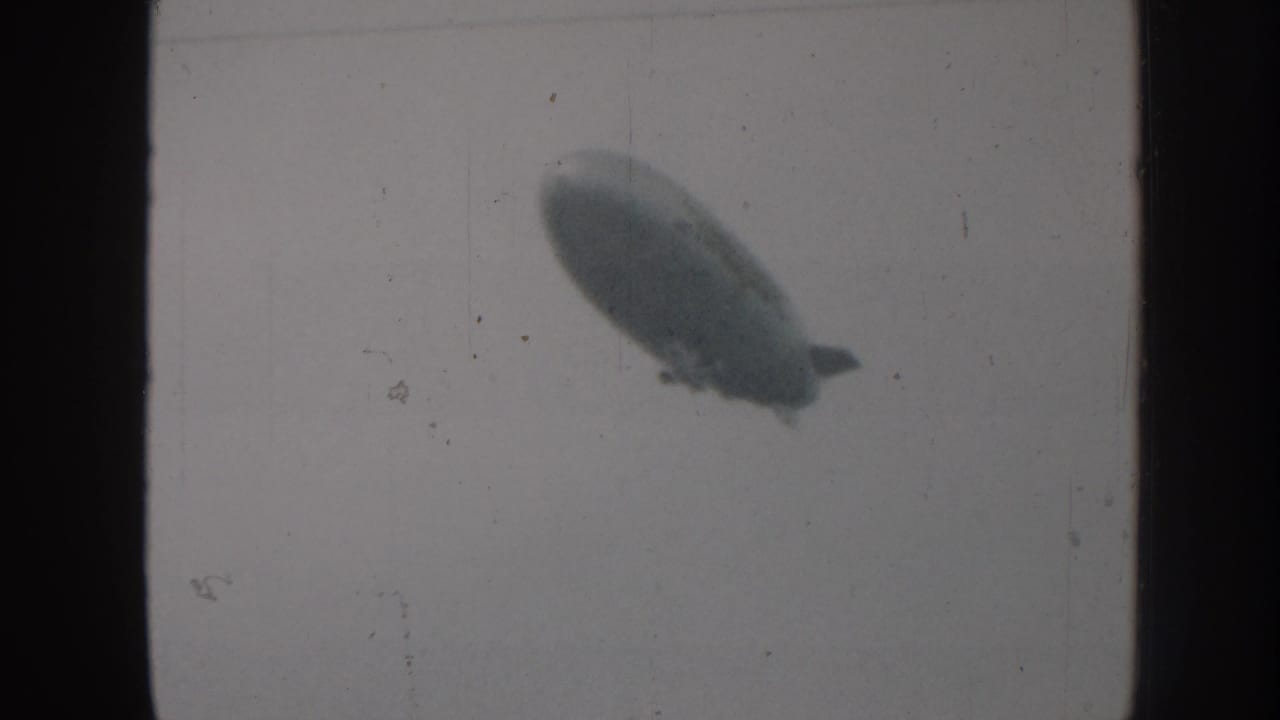 Cigar-shaped blimp traversing overcast sky in 8mm home movie footage, Nebraska USA, circa 1958, with film grain and frame-edge vignetting.