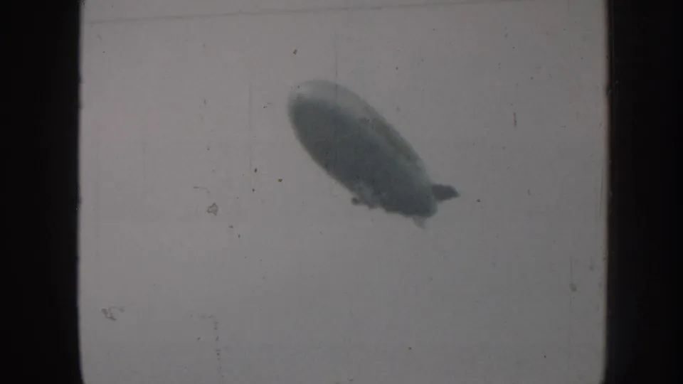 Cigar-shaped blimp traversing overcast sky in 8mm home movie footage, Nebraska USA, circa 1958, with film grain and frame-edge vignetting.