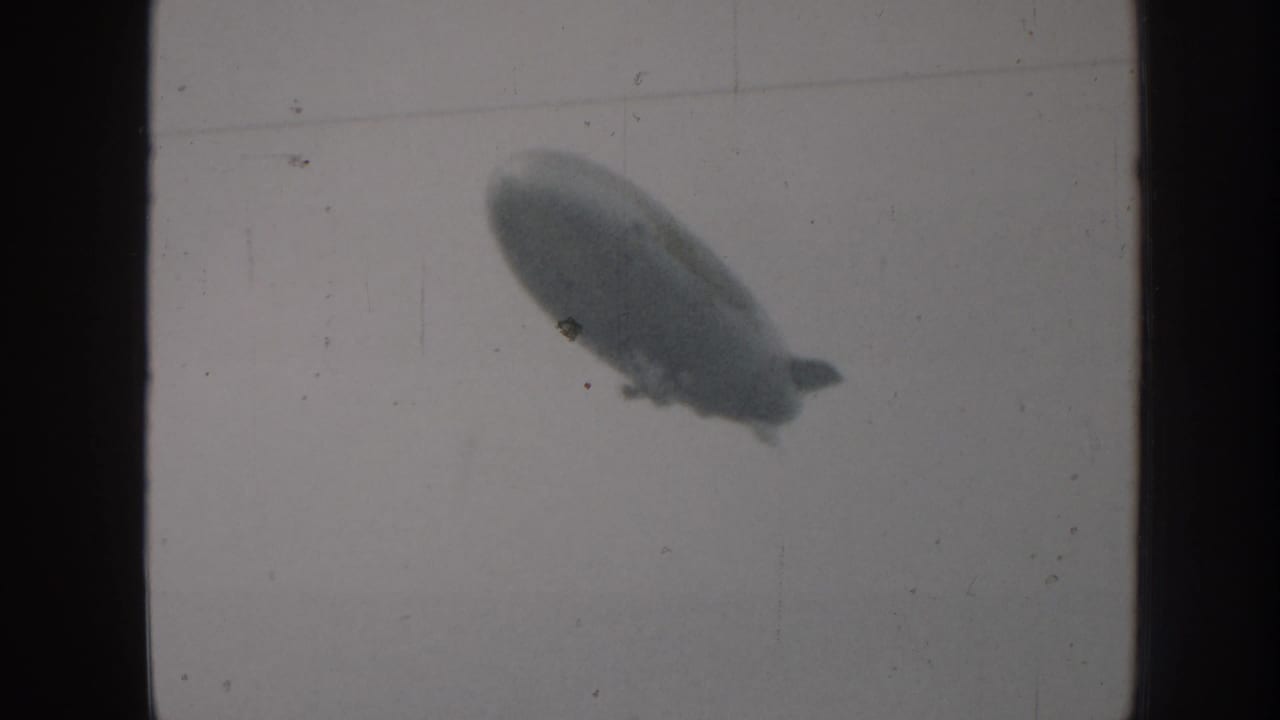 Blimp in profile slightly right of center against a uniform pale grey sky, 8mm grain and dust specks visible.
