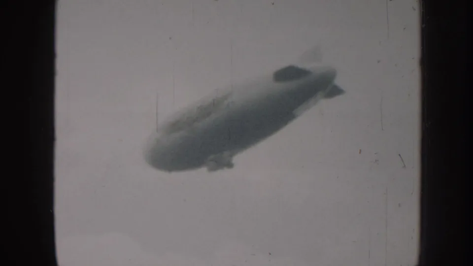 Grainy 8mm monochrome frame, likely Nebraska 1958, of an airship at medium distance with gondola and tail fins visible against pale sky.