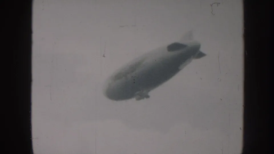 Grainy 8mm monochrome frame, likely Nebraska 1958, showing an airship with prominent cruciform tail fins drifting across an overcast sky.