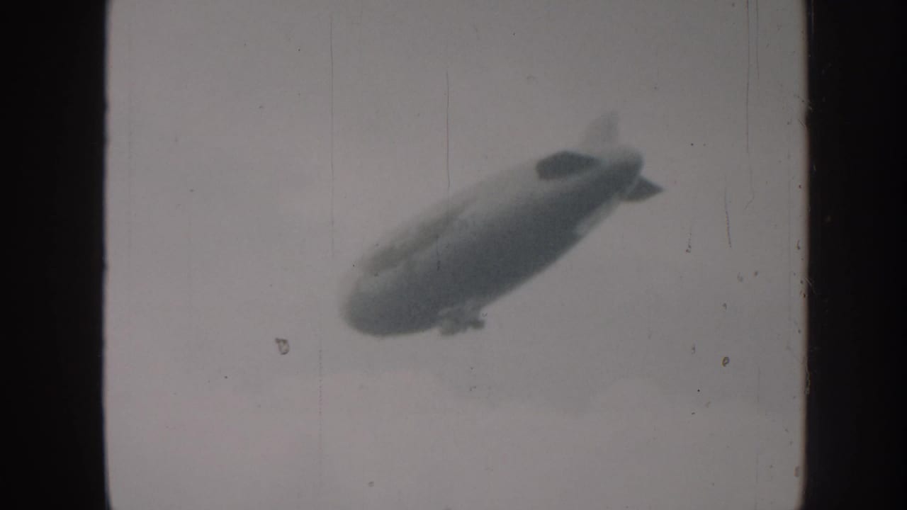 An airship with visible tail fins and gondola tracks upper-left across a hazy sky in 8mm home-movie footage consistent with late-1950s amateur cine practice.